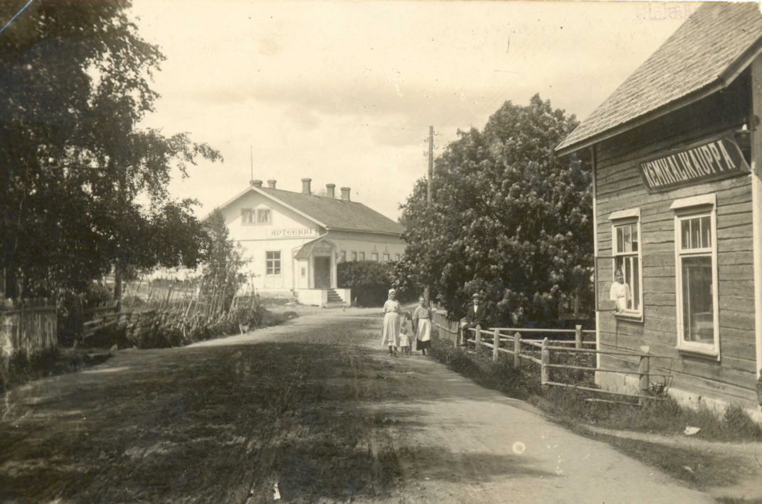 Tie jolla neljä lasta kävelemässä oikealla rakennus jossa teksti Kemikalikauppa ja vasemmalla vaalea rakennus jossa teksti Apteekki.