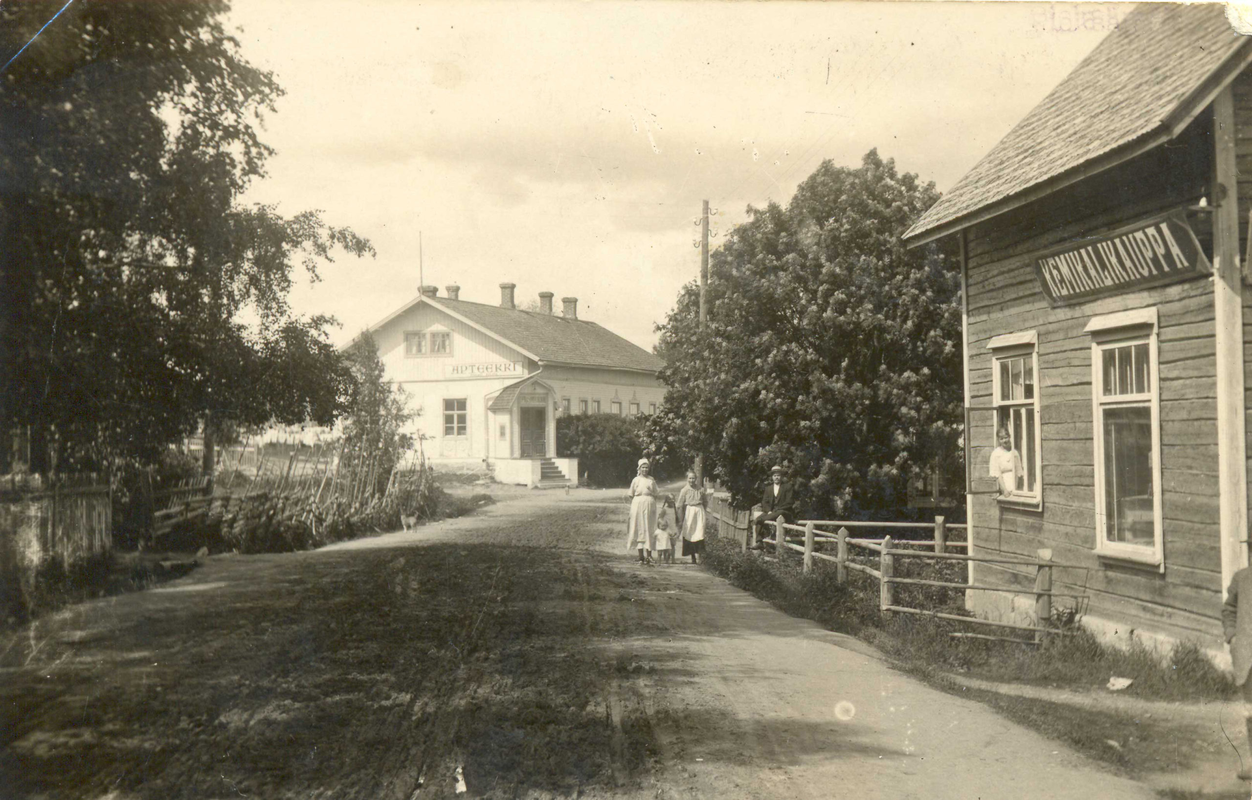 Tie jolla neljä lasta kävelemässä oikealla rakennus jossa teksti Kemikalikauppa ja vasemmalla vaalea rakennus jossa teksti Apteekki.
