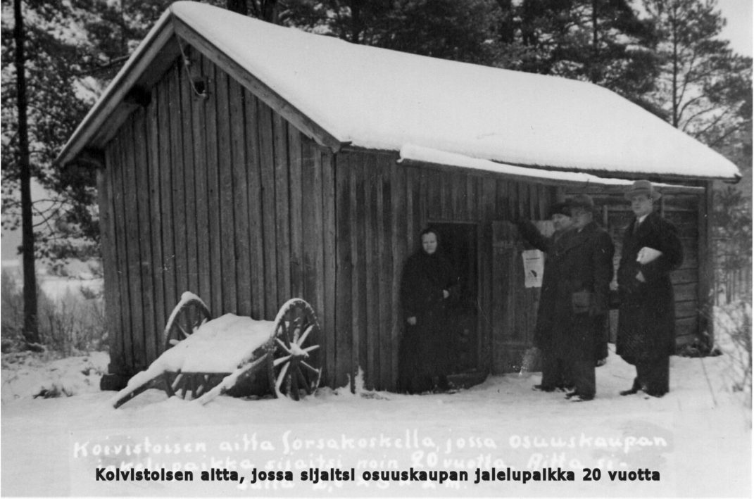 Pieni puinen rakennus jonka ovella nainen mekossa ja seinustalla kolme miestä kuvassa teksti Koivistoisen aitta jossa sijaitsi Osuuskaupan jakelupista 20 vuotta.