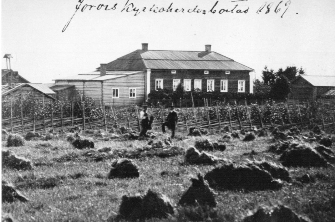 Etualalla heinäpelto jota rajaa aita johon nojailee kaksi miestä taustalla suuri puurakennus jonka päätyyn tehty laajennus kuvaan kirjoitettu teksti Jorois kyrkoherde bostad 1869.