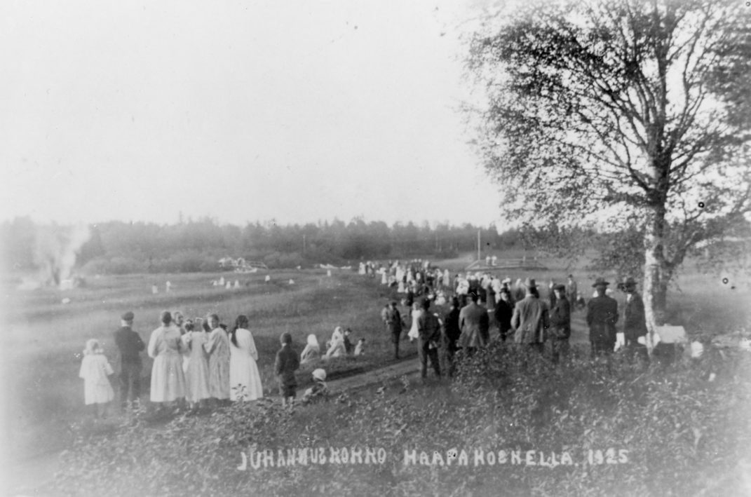Aukea maisema jossa suuri joukko ihmisiä seisomassa katsomassa vasemmassa laidassa olevaa kokkoa kuvassa teksti Juhannuskokko Haapakoskella 1925.