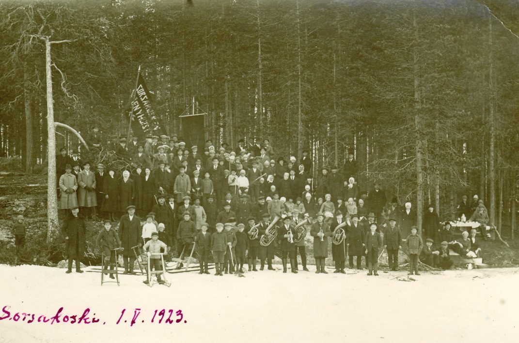 Suuri ihmisjoukko jolla mukana lippuja etualan ihmisillä sylissä puhallinsoittimia ja lisäksi etualalla lapsia potkureilla kuvassa teksti Sorsakoski 1. V. 1923.