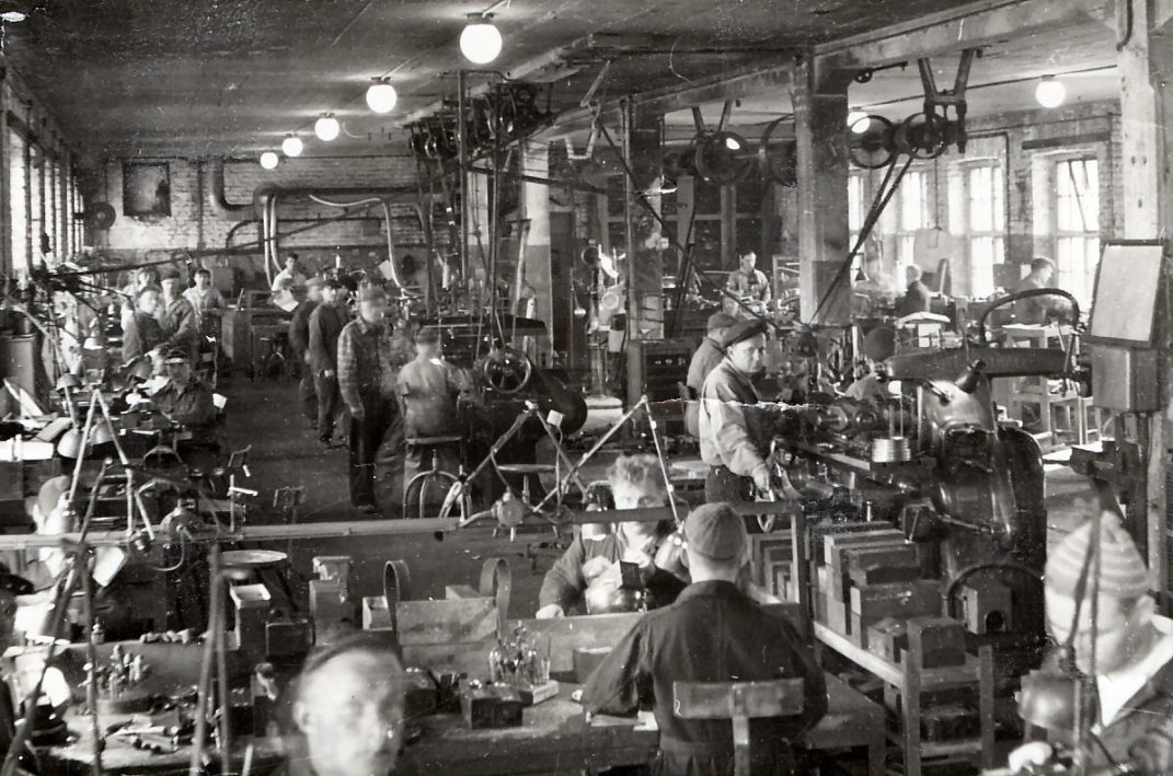 Sisäkuva tehdashallista jossa koneille voimaa tuovia hihnoja ja koneita sekä työntekijöitä.