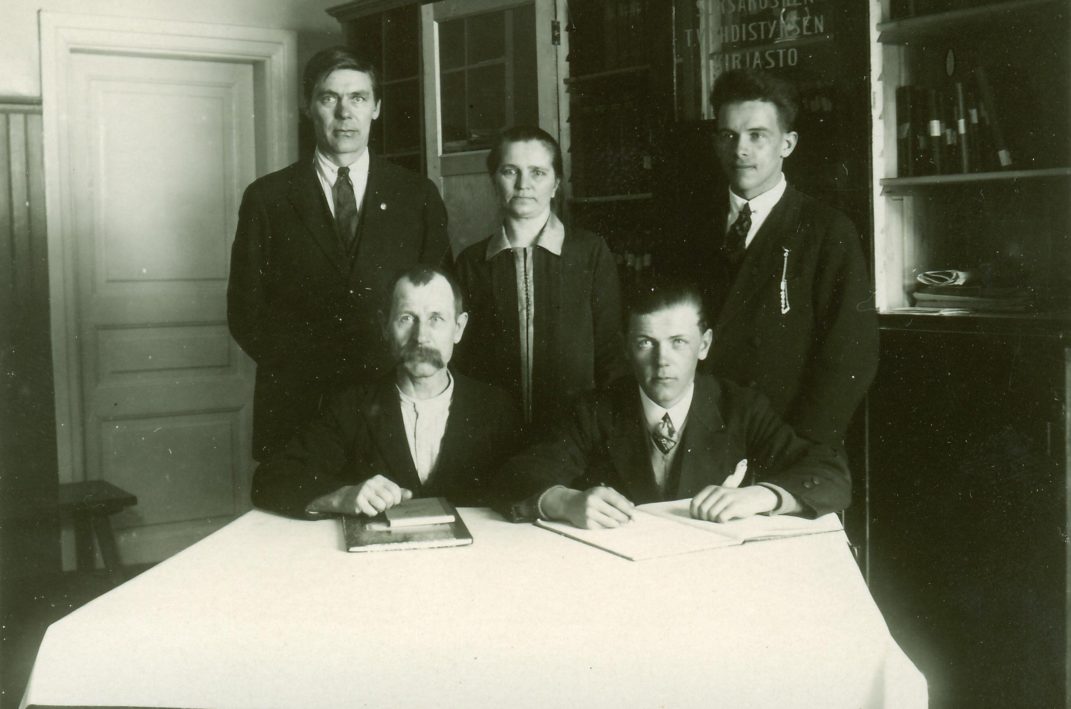 Viisi ihmistä joista kaksi miestä istuu pöydän ääressä ja heidän takana seisoo kaksi miestä ja heidän keskellä yksi nainen.