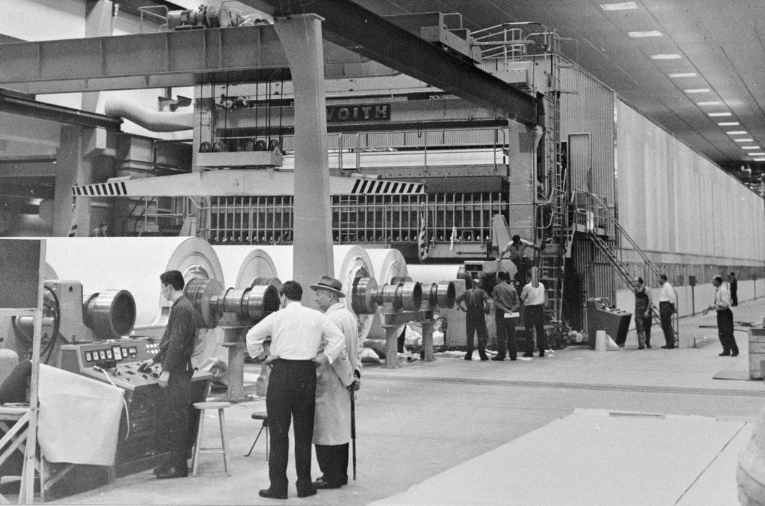 suuri tehdashalli jossa paperikone jonka edustalla seisoskelee ihmisiä