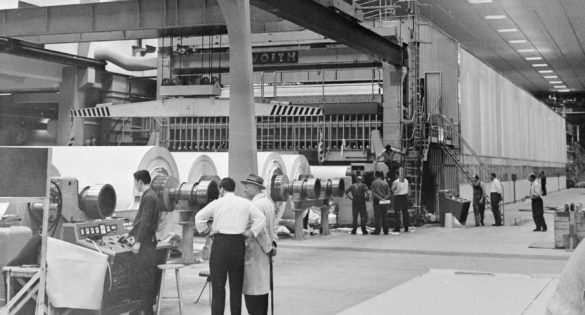 Suuri teollisuushalli jossa paperikone etualalla ihmisiä seisoskelemassa
